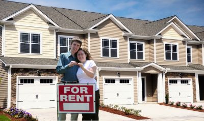 a couple standing behind a for rent sign holding keys with townhomes in the background